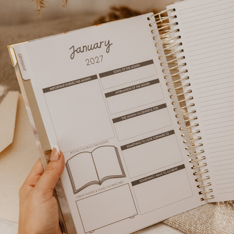 A hand holds an open spiral-bound planner showing the January 2027 page, styled on a neutral fabric surface with warm dried flowers in the background.