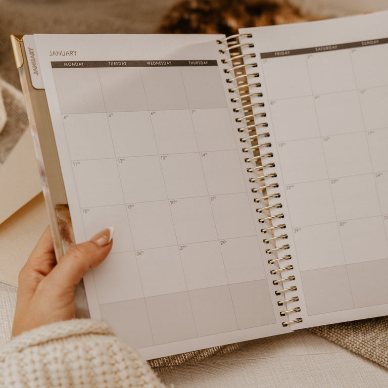 A hand holding an open spiral-bound monthly planner with a minimalist calendar grid, photographed in a warm neutral indoor setting.
