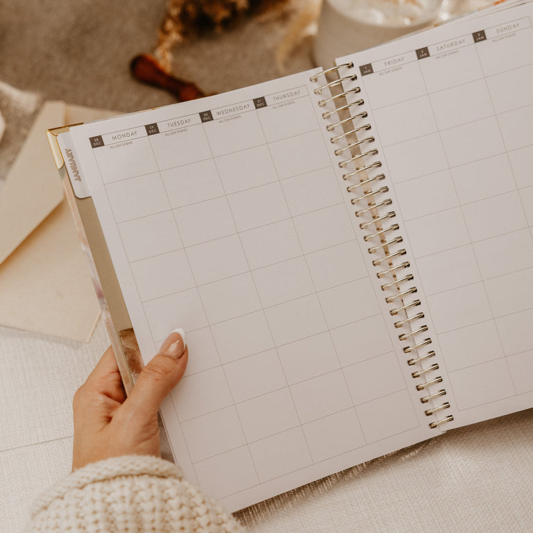 A hand holding an open spiral-bound weekly planner with a clean grid layout, styled on a soft beige fabric surface with dried floral decor in the background.
