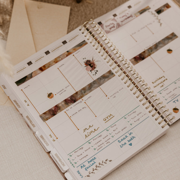 An open spiral-bound planner with weekly and daily layout pages, decorated with floral washi tape and gold accents, styled on a neutral fabric surface with a stamp and envelope nearby.
