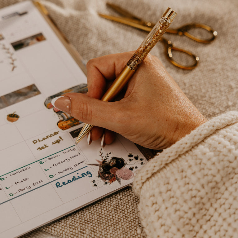 A close-up lifestyle shot of a hand writing in a planner with a gold glitter pen, set on a cozy beige knit blanket with gold scissors and a white mug in the background.