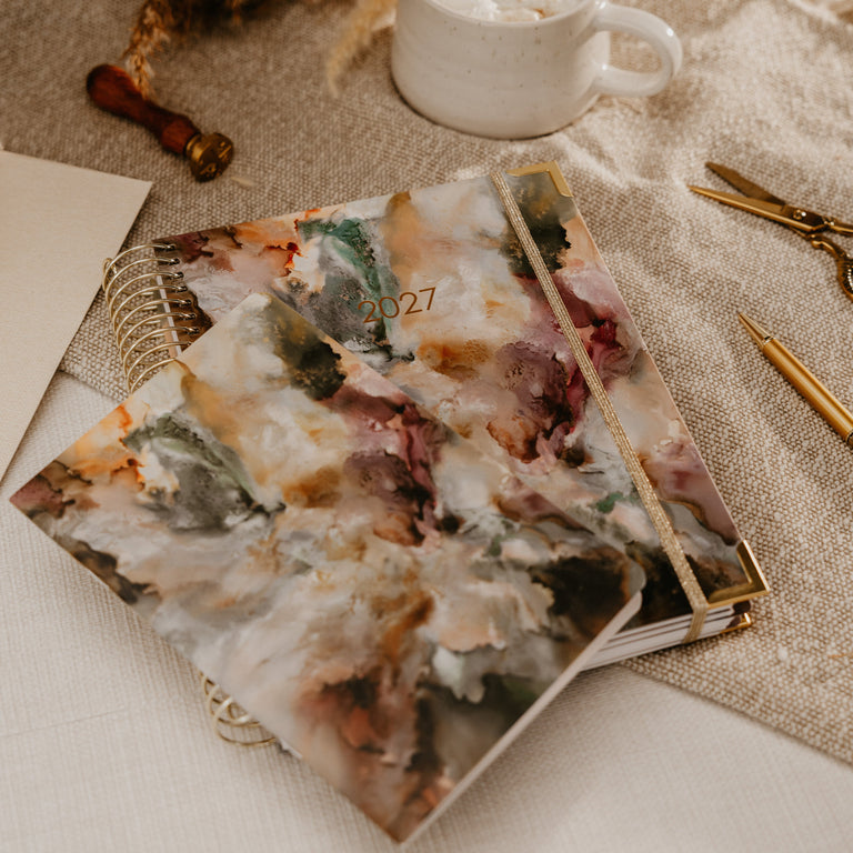 A stack of decorative notebooks with a marbled floral cover design lies on a beige fabric surface beside a white mug, dried grasses, scissors, and gold pens.