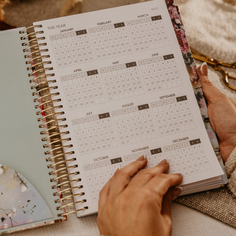 A person's hands holding an open spiral-bound planner showing a monthly calendar page, set against a cozy background with a mug of coffee and neutral-toned fabric.