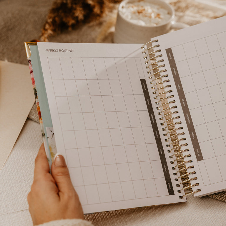 A hand holding an open spiral-bound weekly planner with grid pages, styled on a beige fabric surface with dried grasses and a small cup in the background.