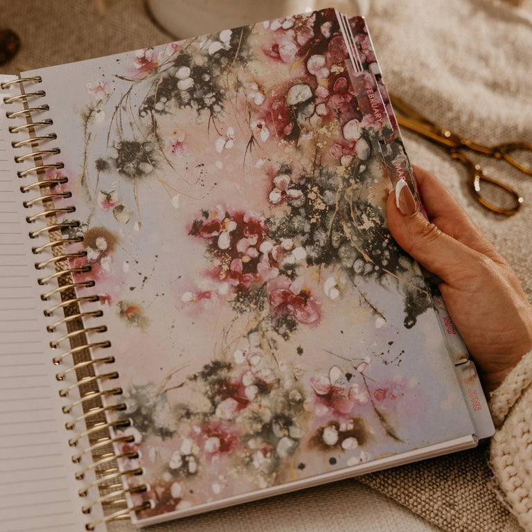 A hand holds an open spiral-bound notebook with a floral cover featuring pink, white, and gray blossoms. The scene is styled on a beige fabric surface with a white mug, dried flowers, and gold scissors in the background.