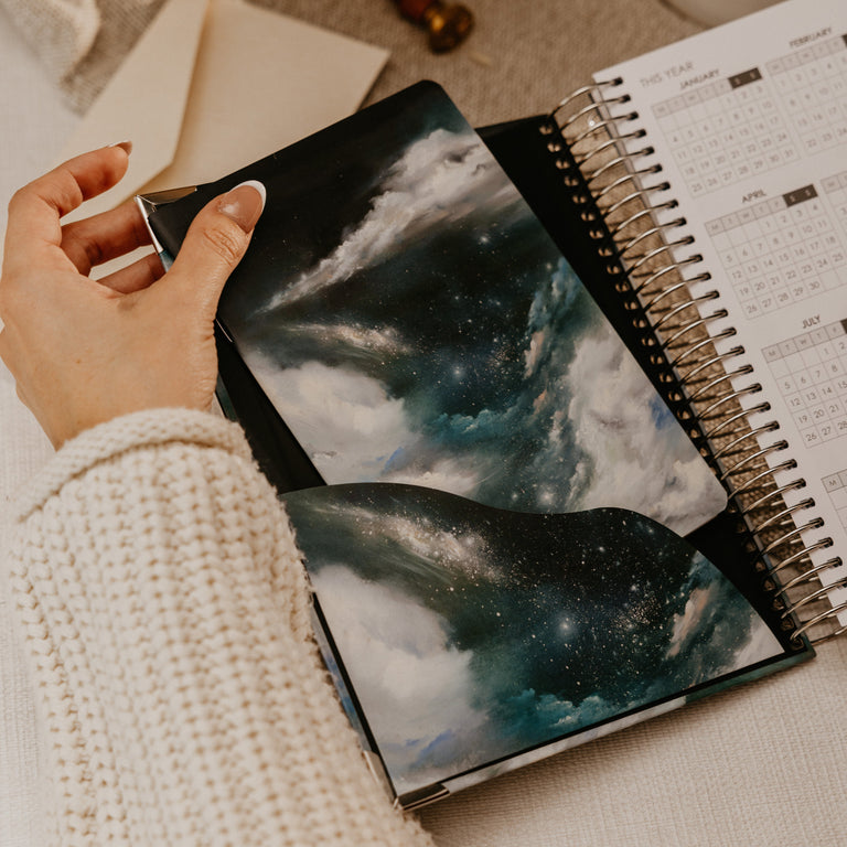 A person wearing a cream knit sweater holds an open spiral-bound planner or journal showing a dark blue and black galaxy cloud cover. The scene is styled on a beige fabric surface with a coffee cup, dried flowers, and a stamp in the background.