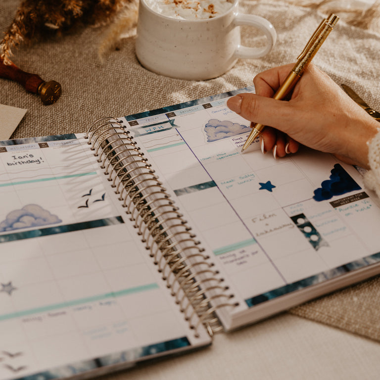 An open spiral-bound planner with decorative blue and white pages, being filled out by a hand holding a gold pen. A white mug of whipped cream sits nearby on a neutral fabric surface with dried botanicals in the background.