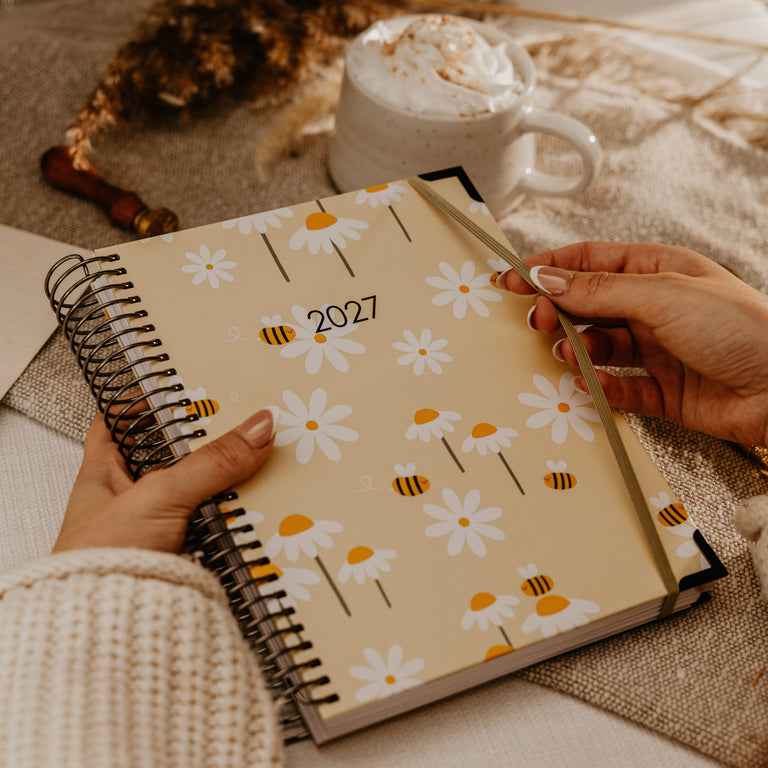 A person holds a spiral-bound 2027 planner with a beige cover decorated with white daisies and yellow-and-black bumblebees. The scene is styled with a white mug of hot chocolate topped with whipped cream and dried grasses on a neutral fabric background.