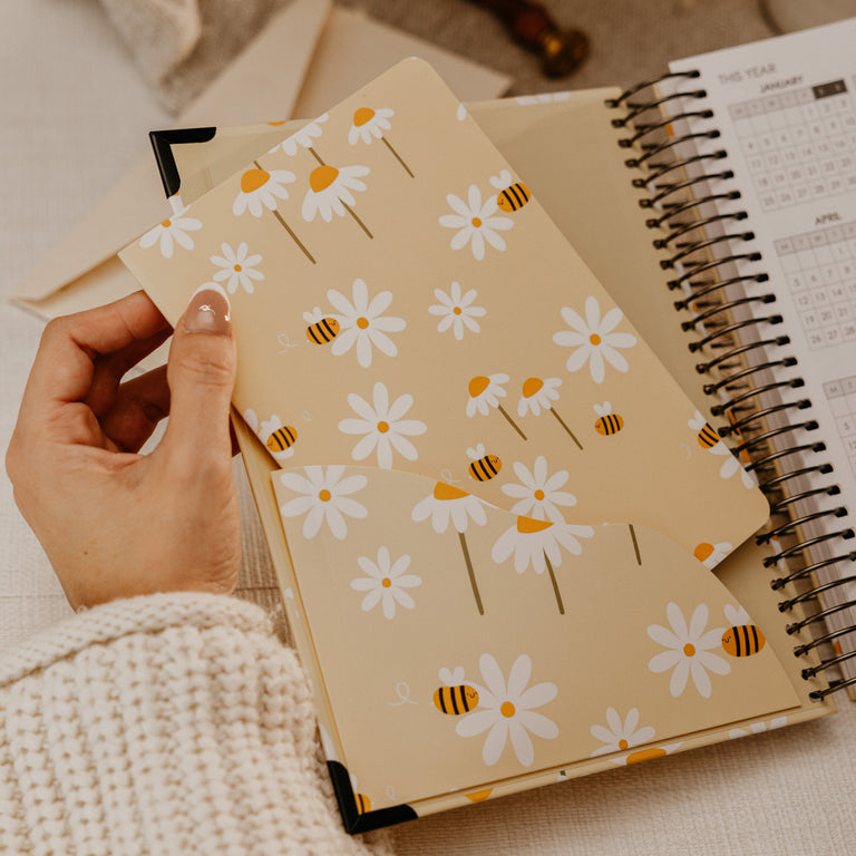 A hand holds a beige pocket organizer with a daisy and bee pattern, layered over an open spiral planner on a neutral fabric surface.