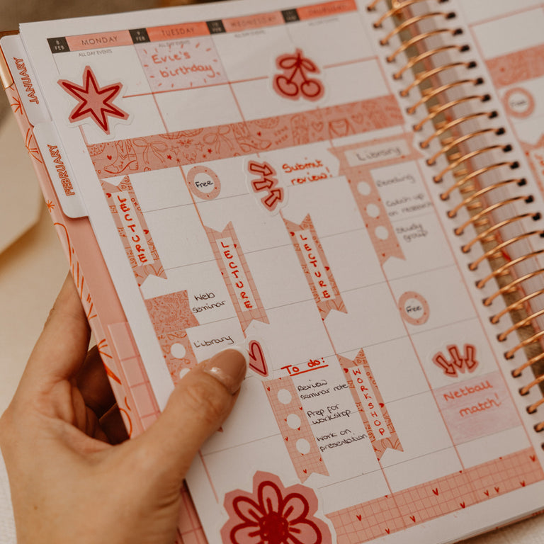 A hand holding an open spiral-bound planner with pink and peach decorative inserts and gold-tone wire binding, photographed in a warm indoor setting.