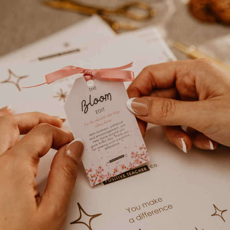 A close-up of hands holding a small printed tag tied with a pink ribbon over a white card. The tag reads 'the bloom 2024' and the card below says 'You make a difference.