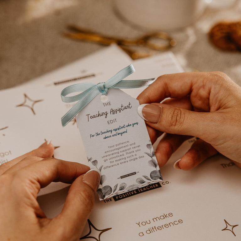 A close-up of hands holding a small gift tag tied with a light blue ribbon over a printed card. The tag reads 'The Teaching Assistant' with elegant script and floral corner details, set against a softly blurred warm tabletop background with dried flowers and gold-toned stationery.