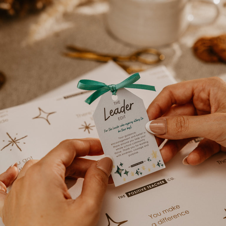 A person's hands holding a white paper tag with a teal ribbon bow. The tag is printed with the text 'The Leader for' and includes small decorative star illustrations.
