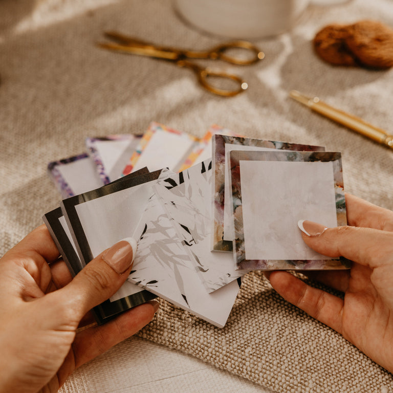A close-up of hands holding a fan of small printed cards or stickers with blank white centers and decorative borders, set on a beige fabric surface with a mug, scissors, and other soft-focus props in the background.