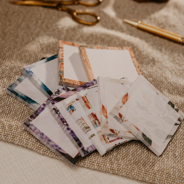 A styled flat-lay of several small patterned sticky notes or cards fanned out on a beige fabric surface, with a white mug, gold scissors, and a gold pen blurred in the background.