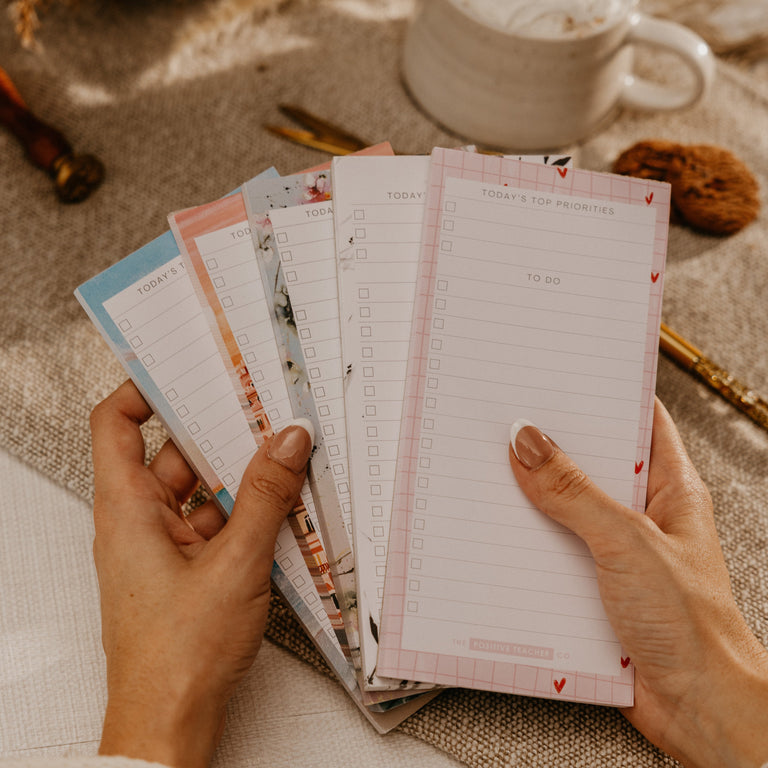 A person's hands holding a fan of lined notepads with pastel covers, set against a cozy background with a cup of coffee, dried flowers, and a cookie.