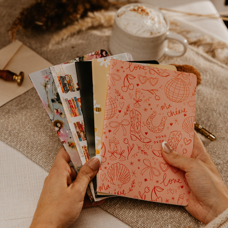 A person holds a fan of colorful patterned cards or envelopes over a cozy beige surface, with a mug of whipped drink and dried grass in the background.