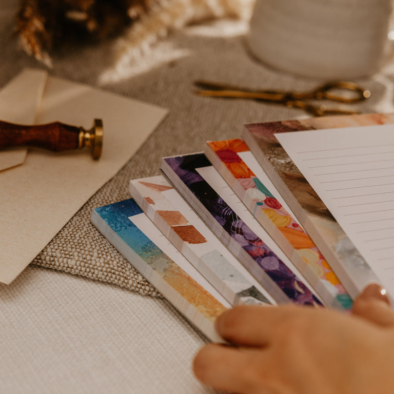 A close-up of a hand holding a fan of colorful patterned planner inserts or bookmarks on a styled desk with a mug, dried flowers, and stationery.