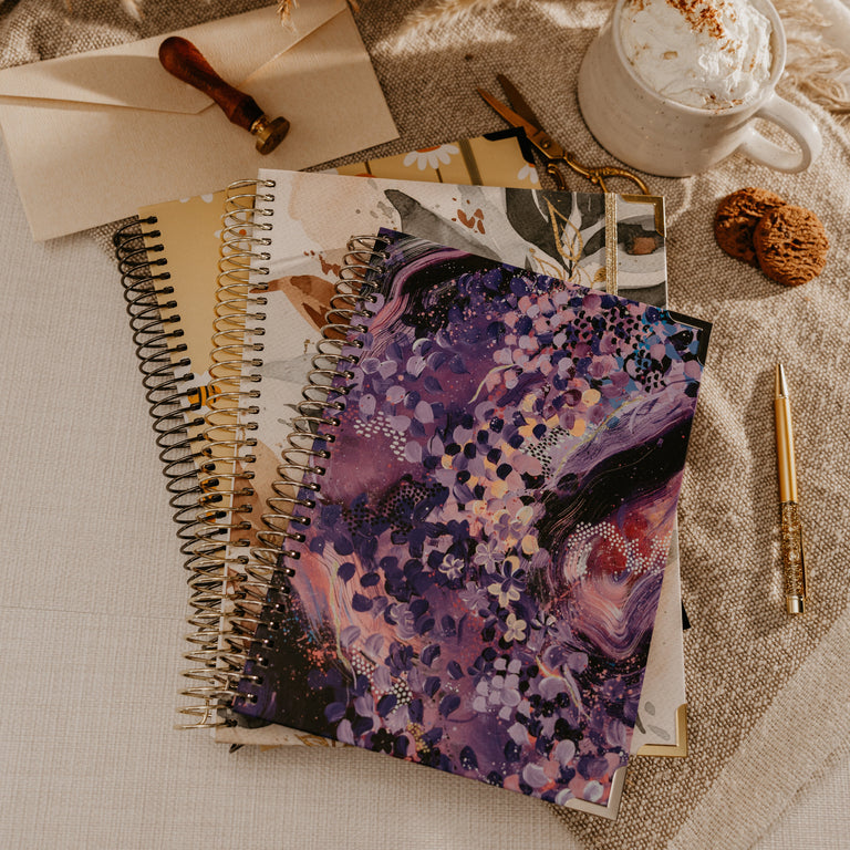 A close-up lifestyle shot of a person's hands holding three spiral-bound notebooks with floral and botanical designs in warm, cozy tones. A ceramic mug of coffee with whipped cream sits in the soft-focus background on a beige fabric surface.
