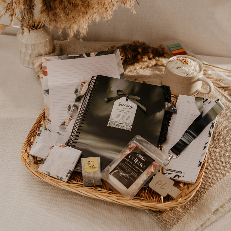 A styled flat-lay of a wicker gift basket filled with stationery and small packaged items, set on a beige fabric surface with dried pampas grass and a mug in the background.