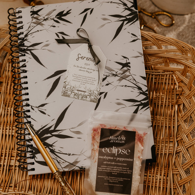 A styled flat-lay of a black and white bamboo-patterned spiral notebook with a ribbon tag, placed on a woven wicker tray alongside a gold pen and a small packet of bath salts labeled 'eclipse'.
