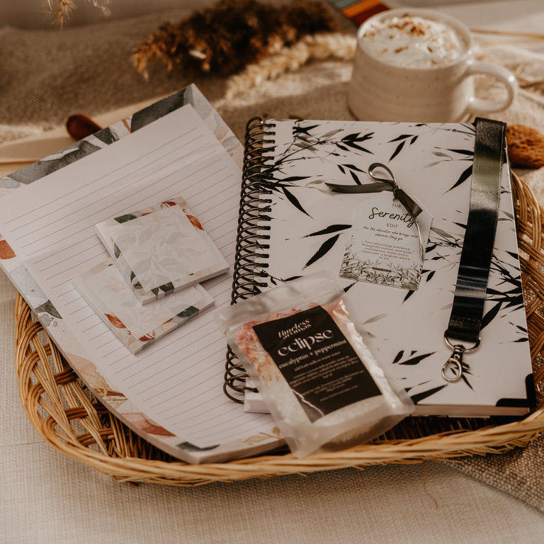 A styled flat-lay of stationery products in a wicker tray, including a black-and-white bamboo print spiral notebook, lined paper, small patterned cards, and a packaged item labeled 'Cupcake' with 'Serenade' branding, set beside a mug of whipped cream with cinnamon.
