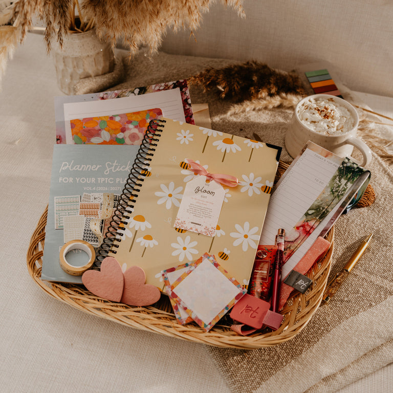 A styled flat-lay of a wicker basket filled with stationery items, including a floral planner, sticky notes, washi tape, pens, and a small cup of hot chocolate, set on a neutral fabric surface with dried grass decor.