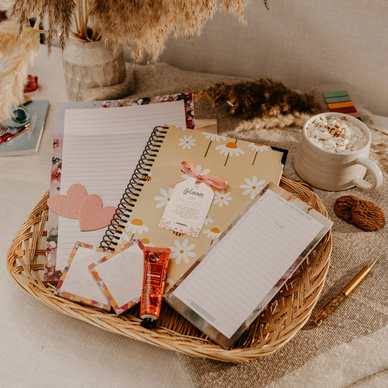 A styled flat-lay of a stationery gift set in a woven basket, featuring a floral planner, notepad, sticky notes, and a candy, arranged on a linen surface with a mug of hot chocolate and dried pampas grass in the background.
