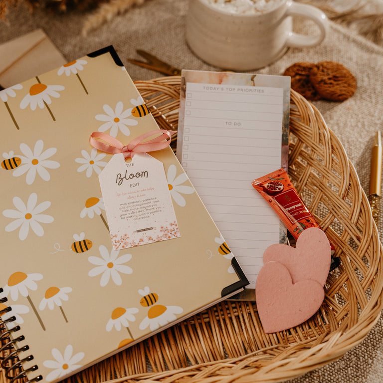 A styled flat-lay featuring a beige spiral notebook with a daisy and bee pattern, tied with a pink ribbon and tag that reads 'Bloom'. The notebook is arranged in a round woven wicker tray alongside a lined notepad, a pink heart-shaped sticker, a small orange item, and a white mug of cappuccino in a cozy, warm setting.