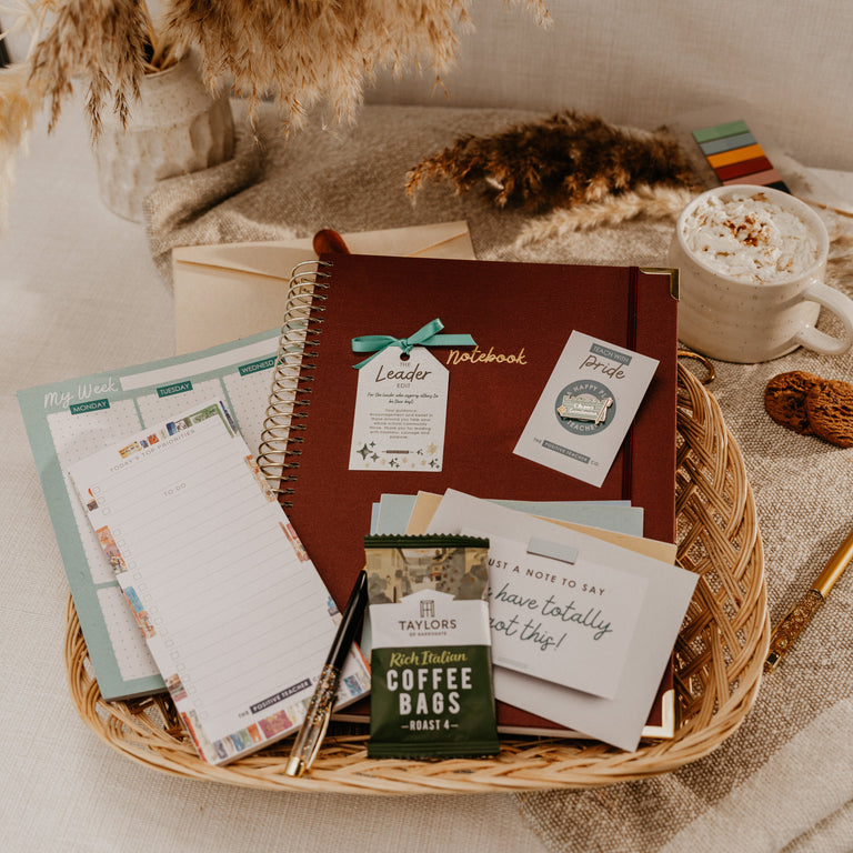 A styled flat-lay of a curated gift basket or bundle in a woven wicker tray, featuring a brown notebook, stationery, a green coffee bag, and a white mug of hot cocoa on a neutral fabric surface.
