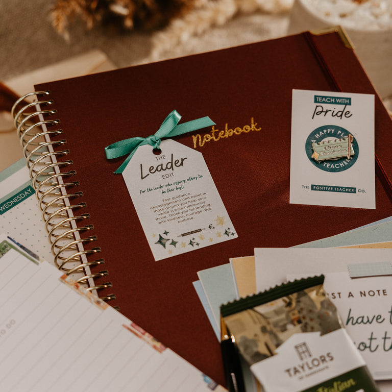 A styled flat-lay of a dark brown spiral-bound notebook with a teal ribbon and printed tags, surrounded by stationery, a coffee bag, and a cup of coffee on a warm neutral background.
