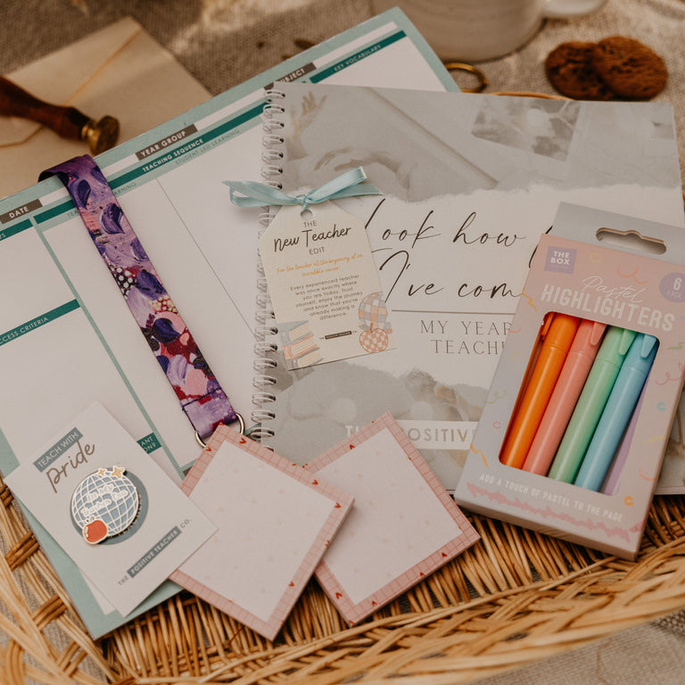 A styled flat-lay of a stationery gift set arranged in a woven basket, featuring a spiral notebook, planner inserts, sticky notes, a floral pen loop, and a box of pastel highlighters, with a coffee cup and dried flowers in the background.