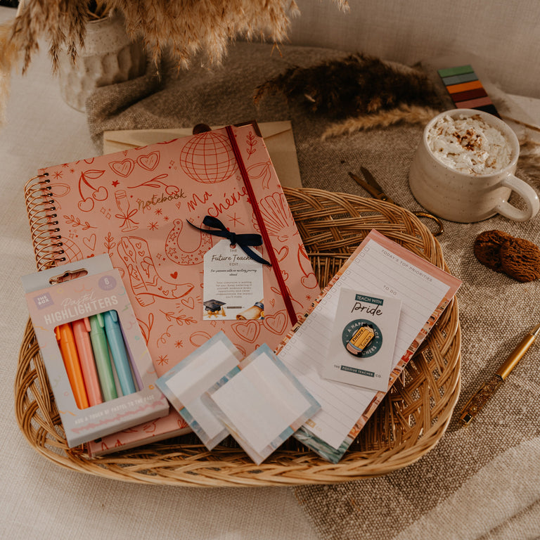A styled flat-lay of a stationery gift set arranged in a woven basket tray, featuring a pink notebook, colorful pens, sticky notes, and notepad cards, set against a cozy neutral background with dried grasses and a mug of coffee.