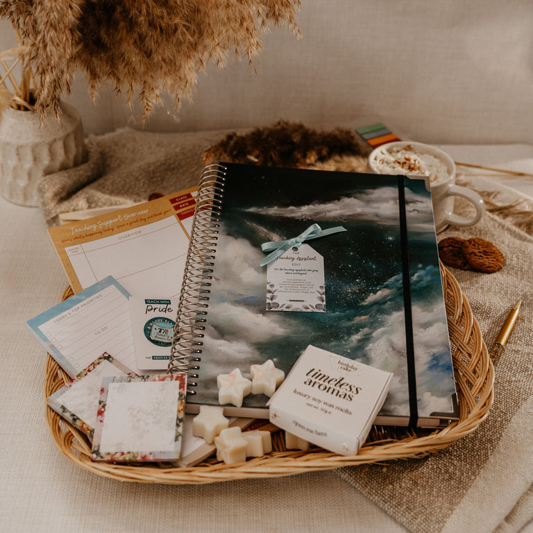 A styled flat-lay of a spiral-bound notebook with a dark blue and gray cloud cover, presented in a wicker tray with small gift cards, star-shaped soaps, and a box labeled 'mindless attention' on a neutral fabric background.