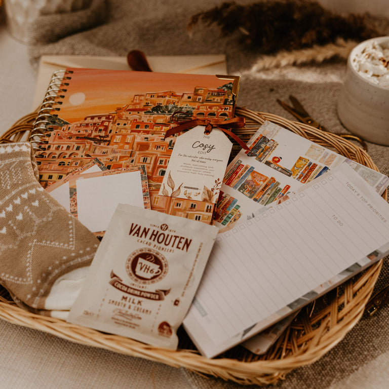 A styled flat-lay of a Van Houten milk chocolate packet placed on top of a wicker basket filled with stationery items, including a notebook, cards, and a small card, set on a neutral fabric surface.
