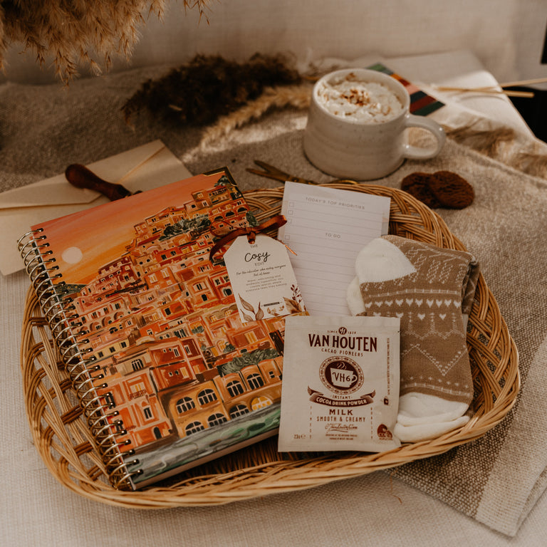 A cozy gift basket arrangement in a woven basket tray, featuring a spiral notebook with a colorful building illustration, a packet of Van Houten milk chocolate, a small card, and folded knit socks, styled with a mug of hot cocoa and dried botanicals on a neutral fabric background.