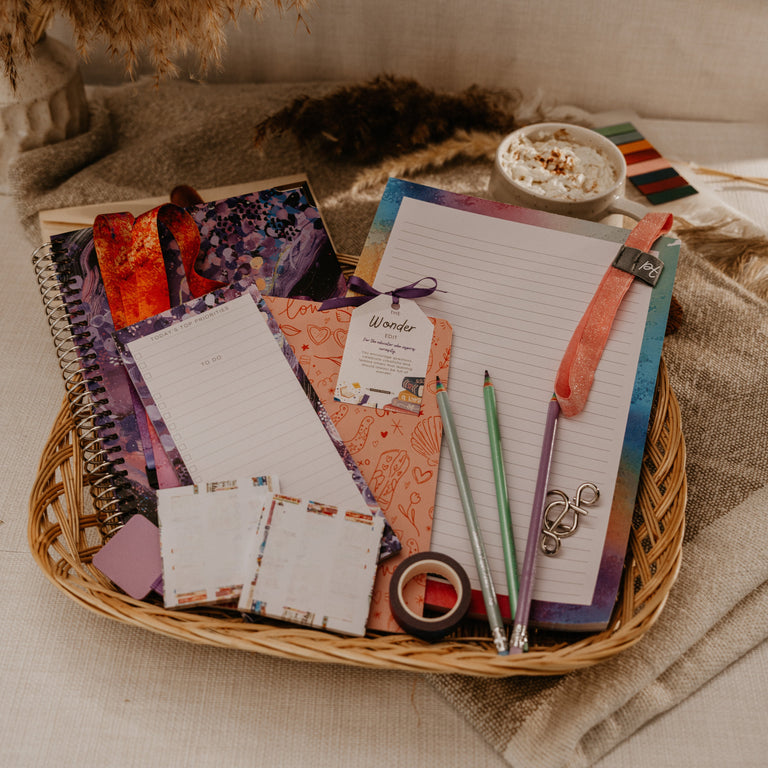 A styled flat-lay of a wicker tray holding a coordinated set of notebooks, notepads, sticky notes, pens, and a small cup of coffee on a beige fabric surface.