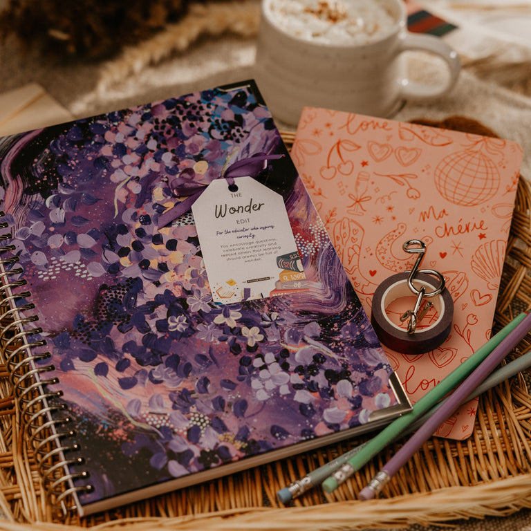 A styled flat-lay of two spiral notebooks in a woven tray, with a mug of whipped cream and dried flowers in the background. The notebook on the left has a purple floral and watercolor cover with a 'Wooden' tag, and the notebook on the right is peach with orange illustrations and the words 'the chase'.