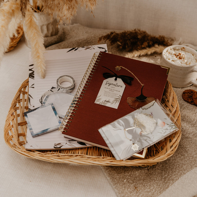 A styled flat-lay of a wicker tray holding a dark red spiral notebook with a 'Reginald' tag, a silver ring, a small silver envelope, and a patterned paper slip, set on a beige fabric surface with dried flowers and a cup of coffee in the background.