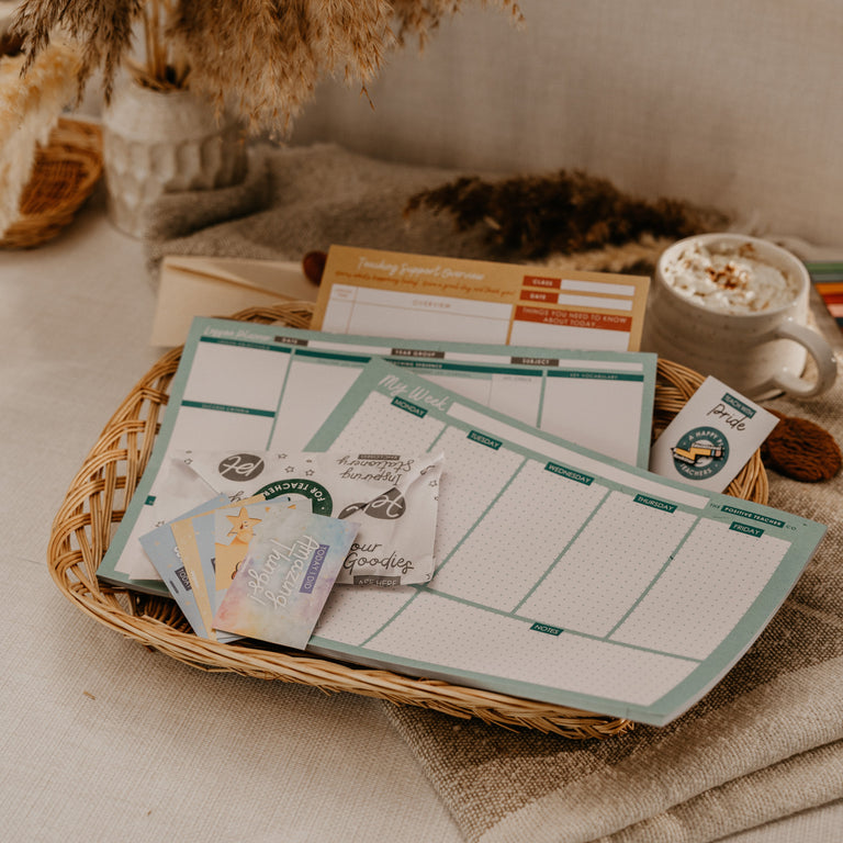 A styled tabletop scene with a wicker basket holding several pastel planner inserts and small cards, surrounded by dried grasses, a mug of coffee, and neutral fabric in the background.