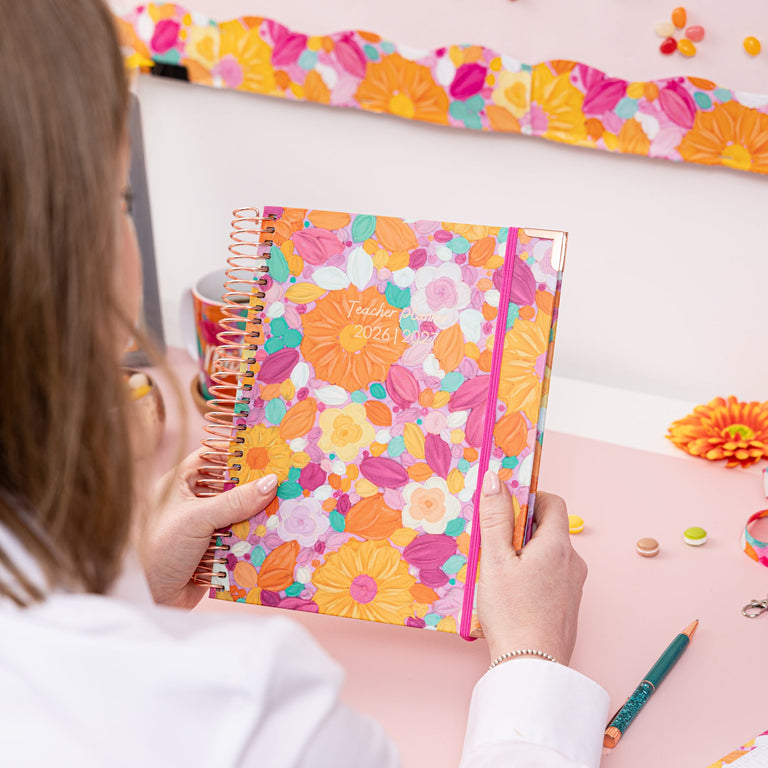 Person holding a colorful notebook with a floral design on a desk with stationery items.