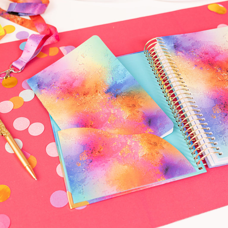 Colorful spiral-bound notebook on a pink surface with decorative ribbons and polka dots.