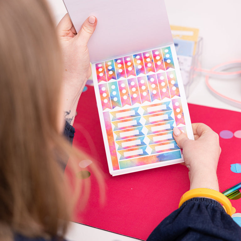 Person holding a colorful planner with stickers on a desk with a rainbow-colored background