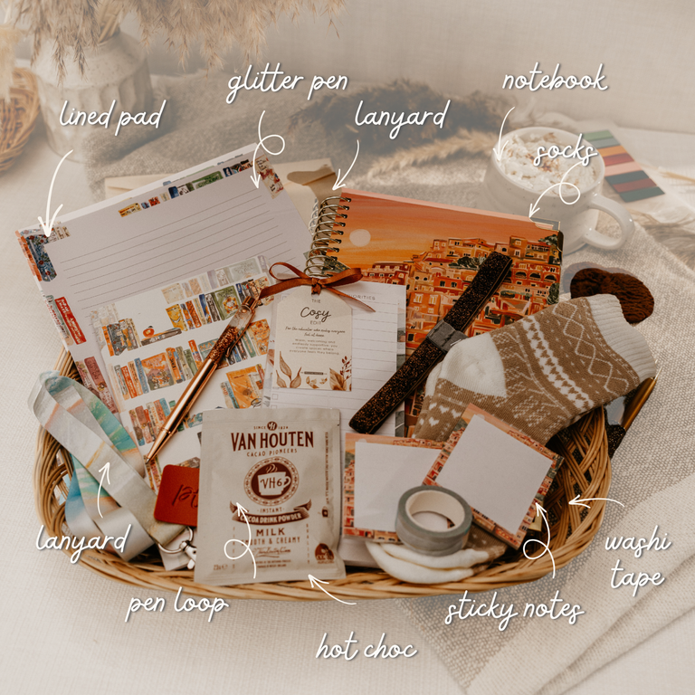 A styled gift basket filled with cozy stationery and sock accessories, including a lined pad, glitter pen, notebook, socks, sticky notes, washi tape, and a Van Houten hot choc packet, arranged on a neutral fabric background with handwritten callouts pointing to each item.