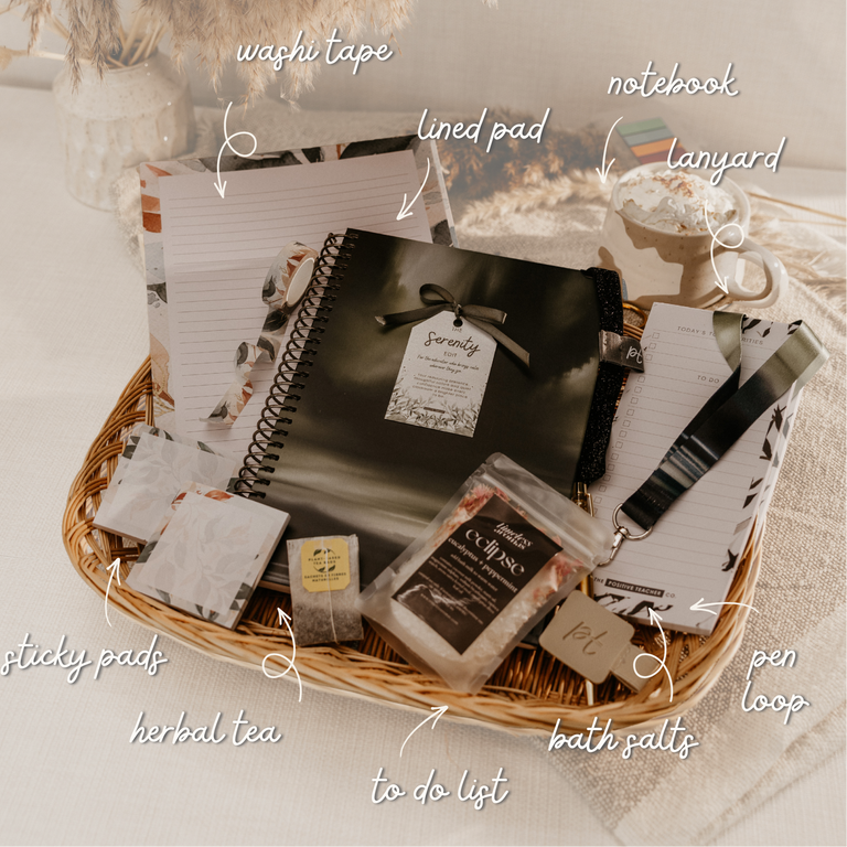 A curated gift basket filled with stationery and self-care items, including a black spiral notebook, lined pads, sticky notes, a pen loop, herbal tea, bath salts, and a notebook lanyard, arranged in a wicker tray on a soft neutral background with handwritten callout labels.